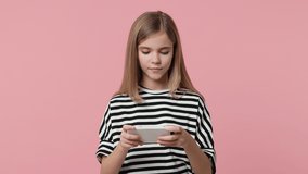 Cheerful excited little kid girl 12-13 years old in basic striped t-shirt isolated on pink background studio. Childhood lifestyle concept. Play game on mobile cell phone doing winner gesture say yes