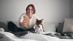 An adult cheerful woman is taking a video-call with her smartphone while sitting on the bed with her basenji dog. 