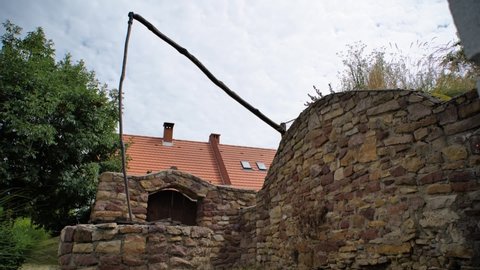 Old Shadoof in the yard of a Hungarian village house. Also known as: sweep-pole well. Traditional fountain construction. Village atmosphere and stone fence. Trees and tiled roof. Rural environment.