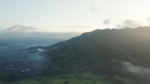 Drone Flight Arcing Over Misty Landscape Of Fields , Village And Tree Covered Hills At Sunrise, Indonesia