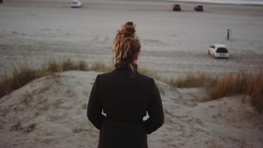 Wide Handheld Tracking Slow Motion Shot Of Young Teenage Girl With Red Hair Tied Up And In Black Coat Walking Away On Sand Dunes On Windy Beach | Shutterstock HD Video #1065870814