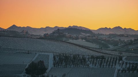 Time lapse sunset to night: famous Barolo wineyards, medieval village perched on hill top, illuminated castle at dusk, snow capped Alps on the background, winter in Italy
