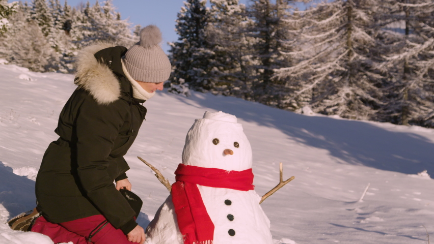 SLOW MOTION, CLOSE UP, PORTRAIT: Happy woman smiles after putting a black hat on a funny snowman she built in an empty snowy meadow. Caucasian female is having fun making a snowman on sunny winter day Royalty-Free Stock Footage #1065871417