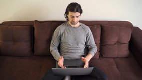 Work from home, home office. A young guy in casual clothes is using laptop for online communication. He sitting on sofa, looks at the laptop screen and speaks 