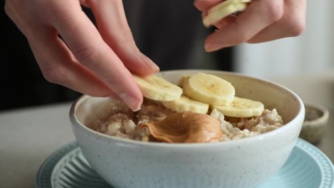 Woman's hands adding banana slices into bowl of oatmeal porridge with peanut butter. Healthy vegan breakfast, weight loss, dieting, clean eating concept