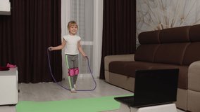 Fit little girl kid doing gymnastics skipping rope workout at home and watching online lessons on laptop pc. Child pupil virtual online distance sport exercises education during coronavirus lockdown