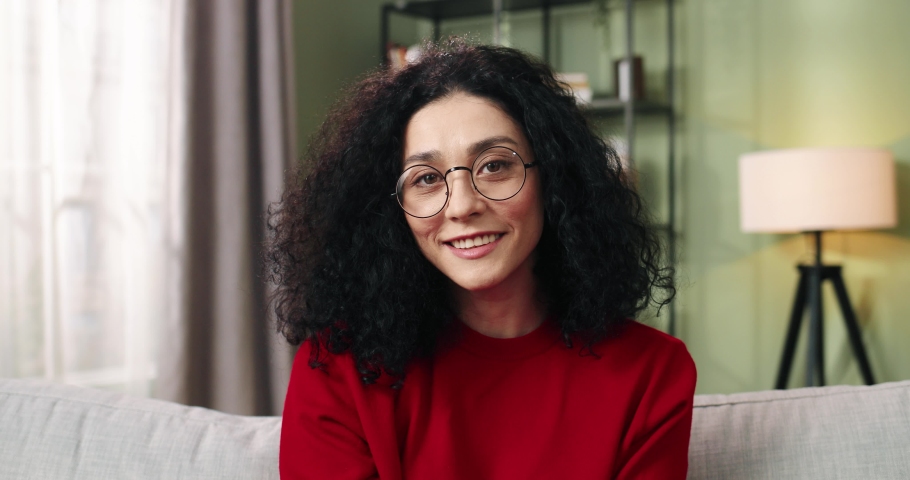 Close up portrait of cheerful happy young millennial woman in glasses sitting at home and looking at came smiling in positive mood. Beautiful Caucasian brunette female in living room. Leisure concept | Shutterstock HD Video #1065879853