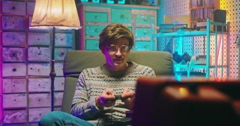 Caucasian man nerd in glasses and with mustache sitting in front of vintage TV monitor and playing videogame with joystick. Male silly goofy loosing in game. Retro style of 80's. Gamer loser from 90's
