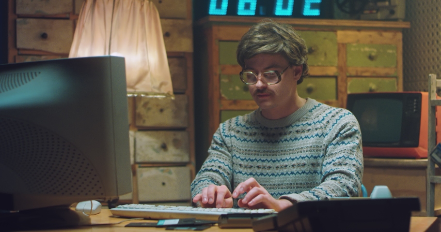 Portrait of Caucasian male goofy nerd in glasses with mustache sitting at desk in retro room and working on computer. Funny man programist looking at camera and typing on keyboard. Vintage style. | Shutterstock HD Video #1065880021