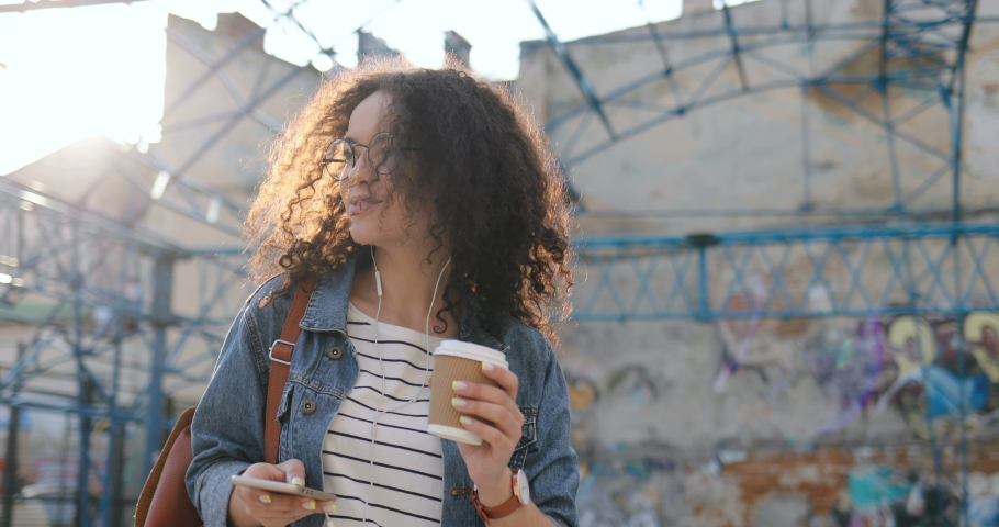 Caucasian good looking young curly woman in glasses tapping on the smartphone while walking outside witha coffee and in headphones.
