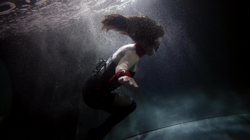 A woman in a black suit floats upside down under the water, her long hair flowing. A mermaid and a nymph. She has a collar around her neck. There are a lot of bubbles floating around. | Shutterstock HD Video #1065891019