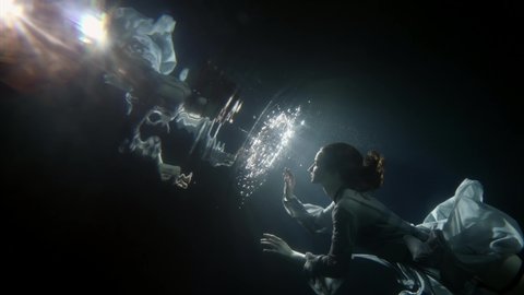 A young woman in a dress floats under the dark water, her hair developing. A mermaid and a nymph. It floats to the surface.