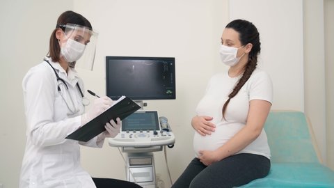 Pregnant woman in medical face mask sitting at gynecological office and signing papers on clipboard that holding doctor. Concept of health care and pandemic time.