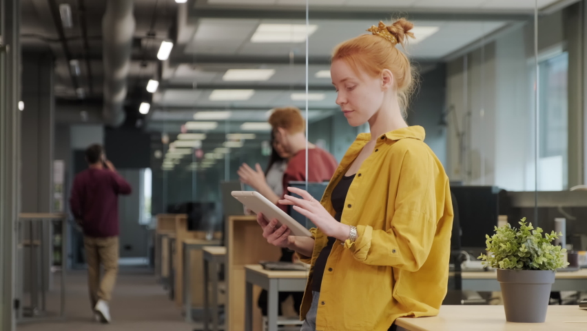 Medium slow-motion shot of young red-haired female office worker in bright yellow shirt working on tablet standing in big spacious office, while coworkers communicating in background | Shutterstock HD Video #1065894664