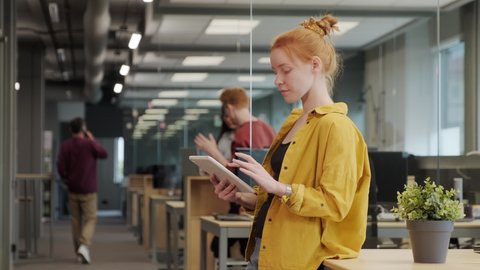 Medium slow-motion shot of young red-haired female office worker in bright yellow shirt working on tablet standing in big spacious office, while coworkers communicating in background