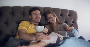 a young couple. a girl and a boy, lying down and relaxing in bed in the bedroom or living room, with a cup of tea in hand, watching a movie, a TV show. Medium shot in slow motion.