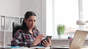 Female watching and uses a cellphone in office. Freelance worker. Focused young woman tapping and scrolling on mobile phone. Close up. Portrait of curlyl brunette girl texting on smartphone, indoor.
