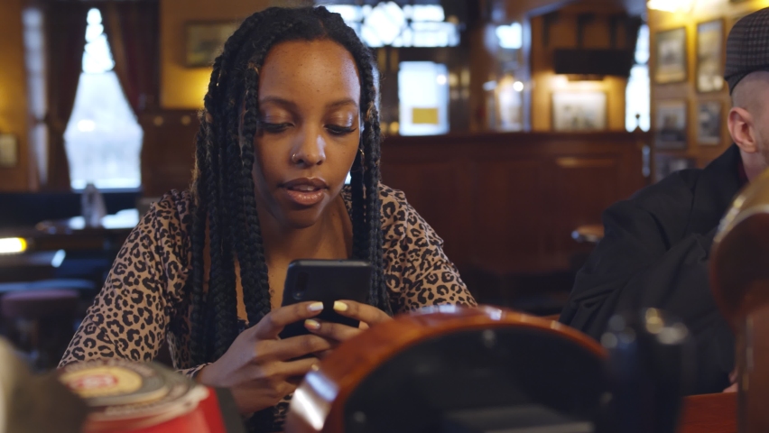 Diverse people sitting at bar counter using smartphone and drinking in pub. Multiethnic young customers relaxing in bar sitting at counter and talking.