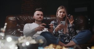 Male female sitting on couch in front of tv screen. Excited millennial man playing video games on console, he loses and gets upset. Woman drinks wine using smartphone. Human relationships concept