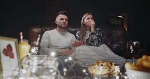 Young couple man and woman watching a horror movie on TV in evening at home, watch scary video in living room at night. Guy scares girl as joke, she takes offense at him and laughs