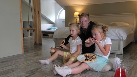 Mom and two daughters are eating pizza, drinking cocktail and watching TV at home sitting on the floor. A family of blondes with freckles are having fun together at home in quarantine.