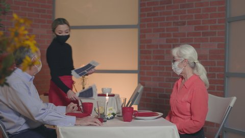 Everyone masked up due to the pandemic this scene tracks past a mature couple sitting in a restaurant who are interacting with a young waitress that hands them each a menu.