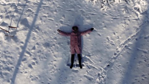 The girl lies in the snow top view. Drone shot. The girl makes an angel on the snow in the winter forest. The drone rises up. Winter park from above.