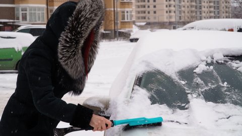 Woman in a Sheepskin Coat with a Hooded Fur Cleans the Car of Snow in Winter in Cold Weather. It is snowing. High quality 4k footage