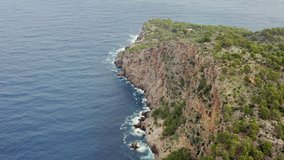 Rocky coastline in the mediterranean sea dron 4K video. Aerial view of mountain ranges with cliffs, peaks and trees crashing against the waves of the sea on the island of Mallorca Spain.