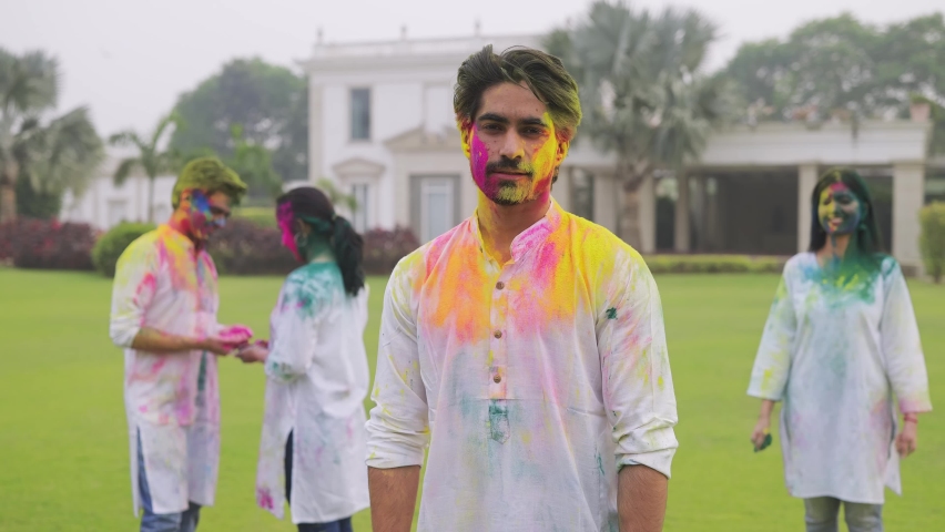 A shot of a young beautiful smiling woman comes from behind and surprises a handsome man by applying color on his face. An attractive Indian couple celebrating Holi festival with friends outdoors. Royalty-Free Stock Footage #1065932341