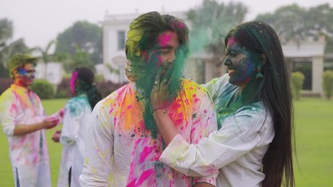 A shot of a young beautiful smiling woman comes from behind and surprises a handsome man by applying color on his face. An attractive Indian couple celebrating Holi festival with friends outdoors.