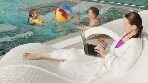 Slow-motion full shot of caucasian family spending time together at roofed thermal pool, woman working on laptop while lying on lounger and father and little girl swimming with beach ball