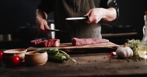 Chef sharpening his knife in front of raw piece of steak and various vegetables. Cooker preparing his tools before cutting raw piece of meat on professional kitchen table, 4k footage