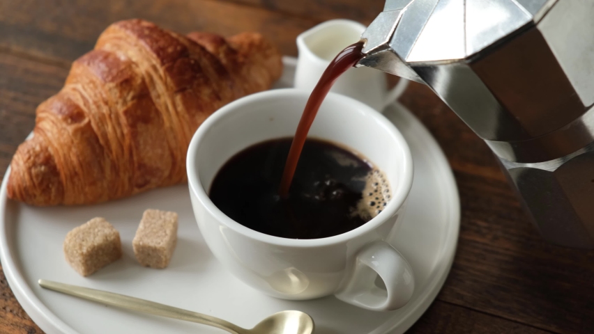 Pouring black coffee in a cup from italian coffee maker Moka pot. Hot black coffee and french croissant served on white plate, wooden table background. Breakfast or lunch in cafe Royalty-Free Stock Footage #1065940414