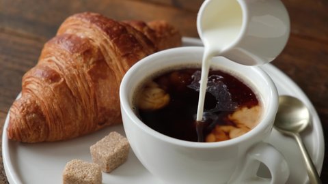 Pouring coffee creamer into cup of black coffee. French croissant and cup of coffee. Tasty sweet breakfast food