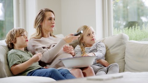 Pregnant mother with children sitting on sofa at home laughing and watching TV with popcorn together - shot in slow motion