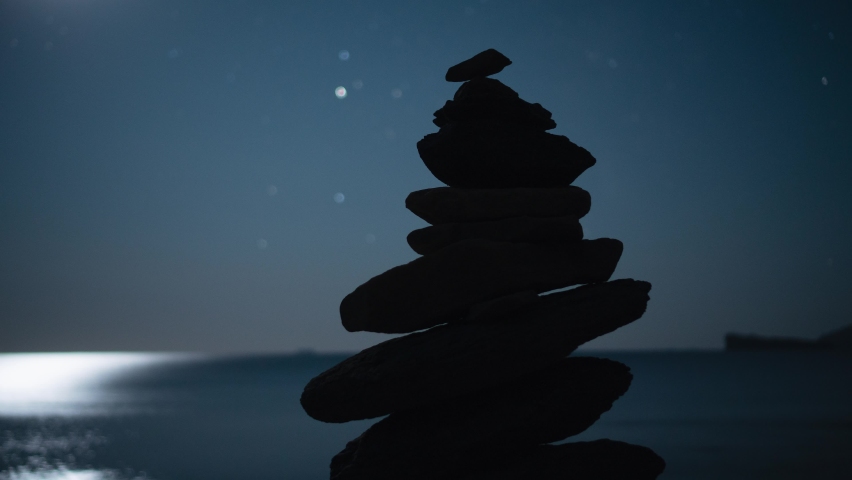 Black pyramid of stones perfect balance against background of moving starry night sky. White bright moon reflected in sea ocean. Coastal natural landscape seascape. Relax. Cinematic details timelapse