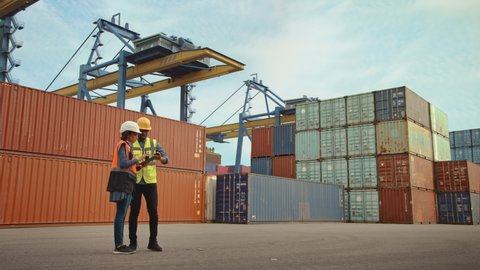 Multiethnic Female Industrial Engineer with Tablet and Black African American Male Supervisor in Hard Hats Stand in Container Terminal. VFX Double Girder Gantry Cranes Work in the Background.