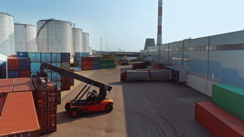 Aerial Drone Footage of a Container Handler Carrying a Large Red Shipping Cargo Container in a Shipyard Terminal. Driver of the Machine is Loading the Crate in the Logistics Center Depot.