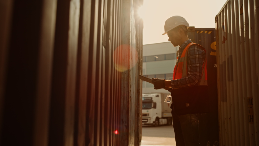 Handsome Caucasian Industrial Inspector or Supervisor Wearing White Hard Hat and High-Visibility Vest is Examining the Containers with His Tablet Computer in a Logistics Shipping Terminal Depot.