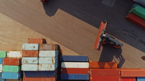 Aerial Drone Footage of a Container Handler Carrying a Large Red Cargo Container in a Shipyard Terminal. Driver of the Machine is Loading the Crate in the Logistics Center Depot. Top Down View.