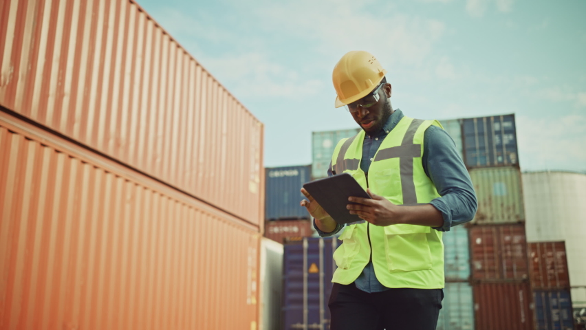 Smiling Portrait of a Handsome African American Black Industrial Engineer in Yellow Hard Hat and Safety Vest Working on Tablet Computer. Inspector or Safety Supervisor in Container Terminal. Royalty-Free Stock Footage #1065943459