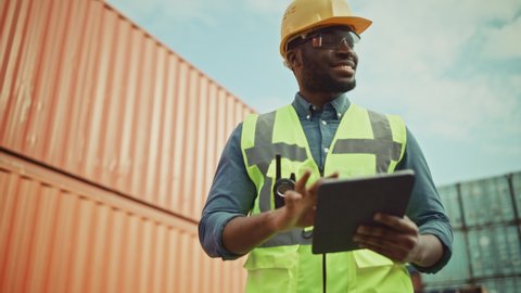 Smiling Portrait of a Handsome African American Black Industrial Engineer in Yellow Hard Hat and Safety Vest Working on Tablet Computer. Inspector or Safety Supervisor in Container Terminal.