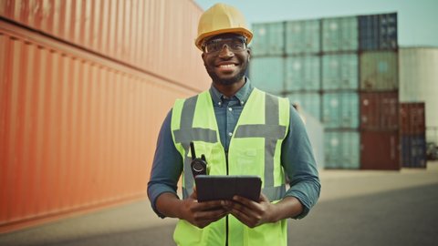 Smiling Portrait of a Handsome African American Black Industrial Engineer in Yellow Hard Hat and Safety Vest Working on Tablet Computer. Foreman or Supervisor in Container Terminal.