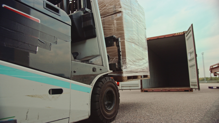 Forklift Driver Loading a Shipping Cargo Container with a Full Pallet with Carboard Boxes in Logistics Operations Port Terminal. Tracking Shot from the Side Shows How Pallet is Brought by the Machine. Royalty-Free Stock Footage #1065943525