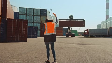 Female Industrial Engineer with Two-Way Radio Wearing White Hard Hat and Orange High-Visibility Vest Pointing the Unloading Location for the Container Handler in Logistics Operation Terminal.