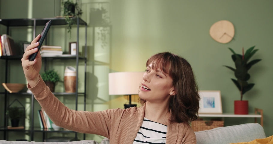 Close up of Caucasian pretty young cheerful female with short haircut touching her hair and posing to smartphone camera while sitting at home in room taking selfie photos on cellphone. Fun concept