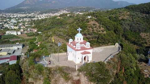 The drone rotates around a greek church on a hill with flags waving and two women standing in front of the church with some houses mountains in the background in Samos Greece Aerial Drone Footage 4K