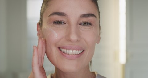 Beauty shot of mature woman applying moisturizing anti wrinkle day or night cream on her previously cleaned face and smiling in camera on golden background. Concept of skincare, cosmetics, healthcare.