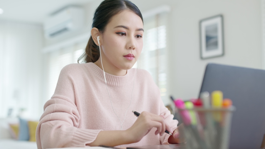 Candid young attractive asian female employee wear earphone listen to digital online course development program in quarantine social distance at home reskill upskill school class for career path. | Shutterstock HD Video #1065950920